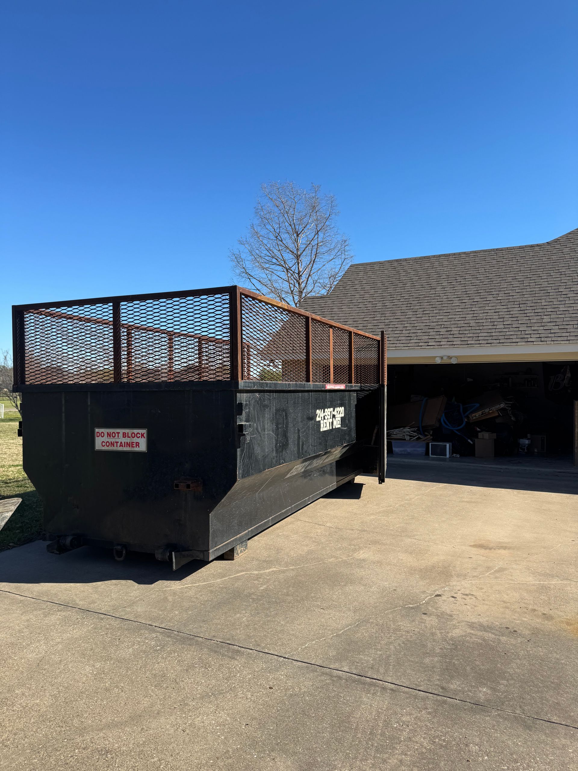 A large black dumpster is parked in a driveway in front of a Frisco house.