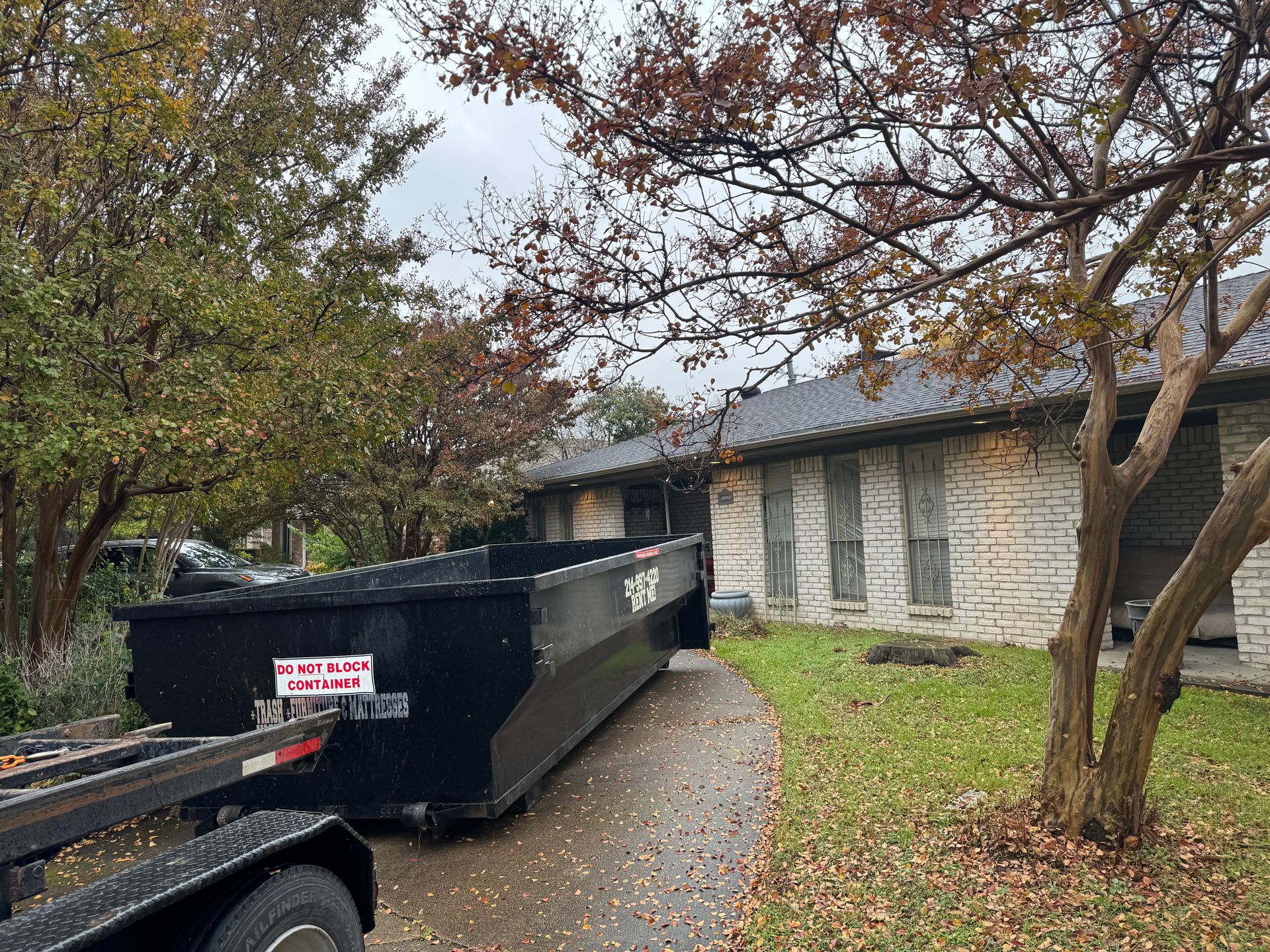 A large black dumpster is sitting in the grass in front of a house.