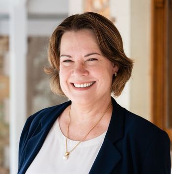 A woman in a blue jacket and white shirt is standing in front of a stone wall.