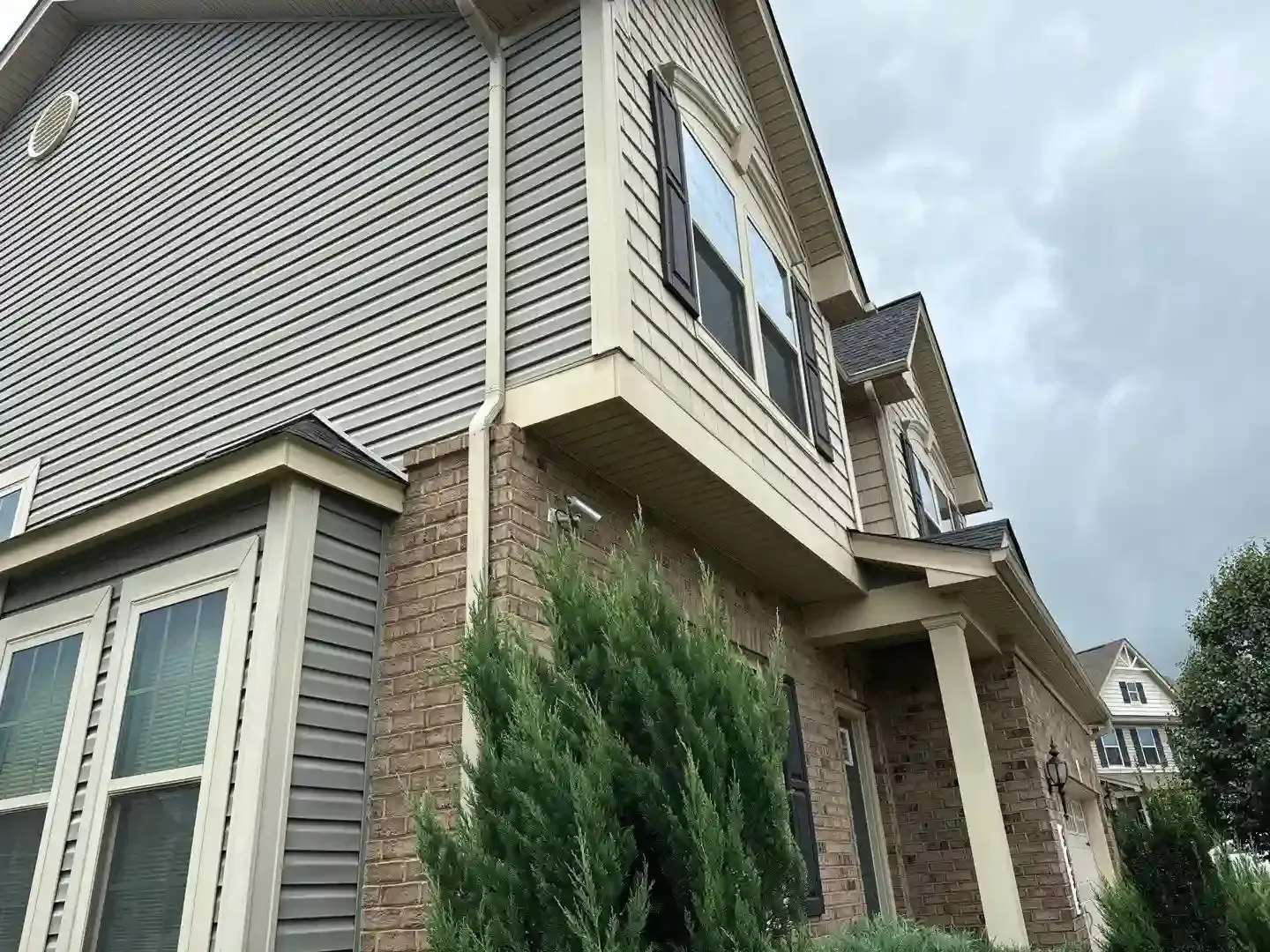 Suburban house exterior with beige siding, brick accents, and green shrubs under a cloudy sky