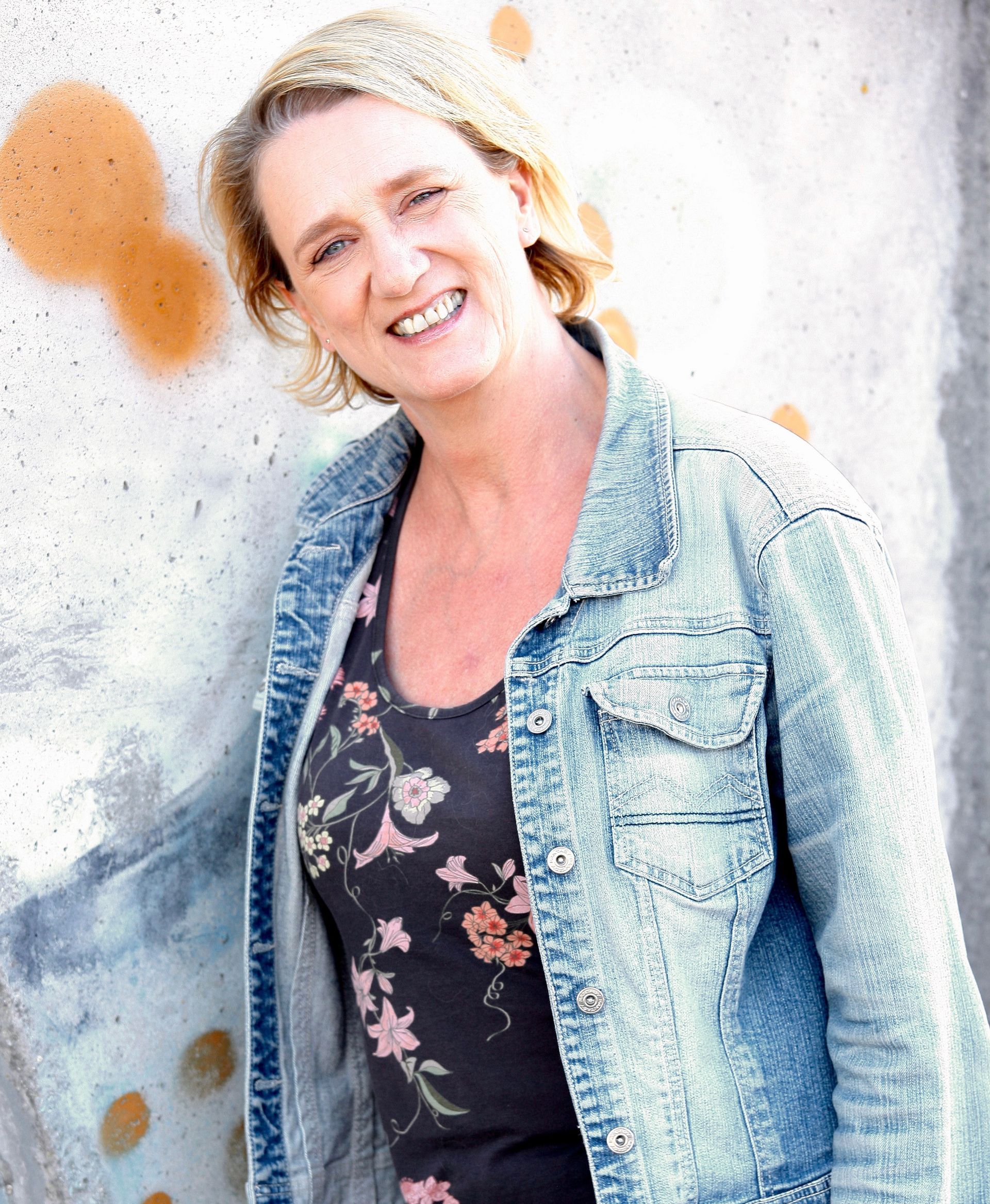 Een lachende Bibian, gekleed in een spijkerjack en een top met bloemenmotief, leunt tegen een met graffiti bedekte muur.