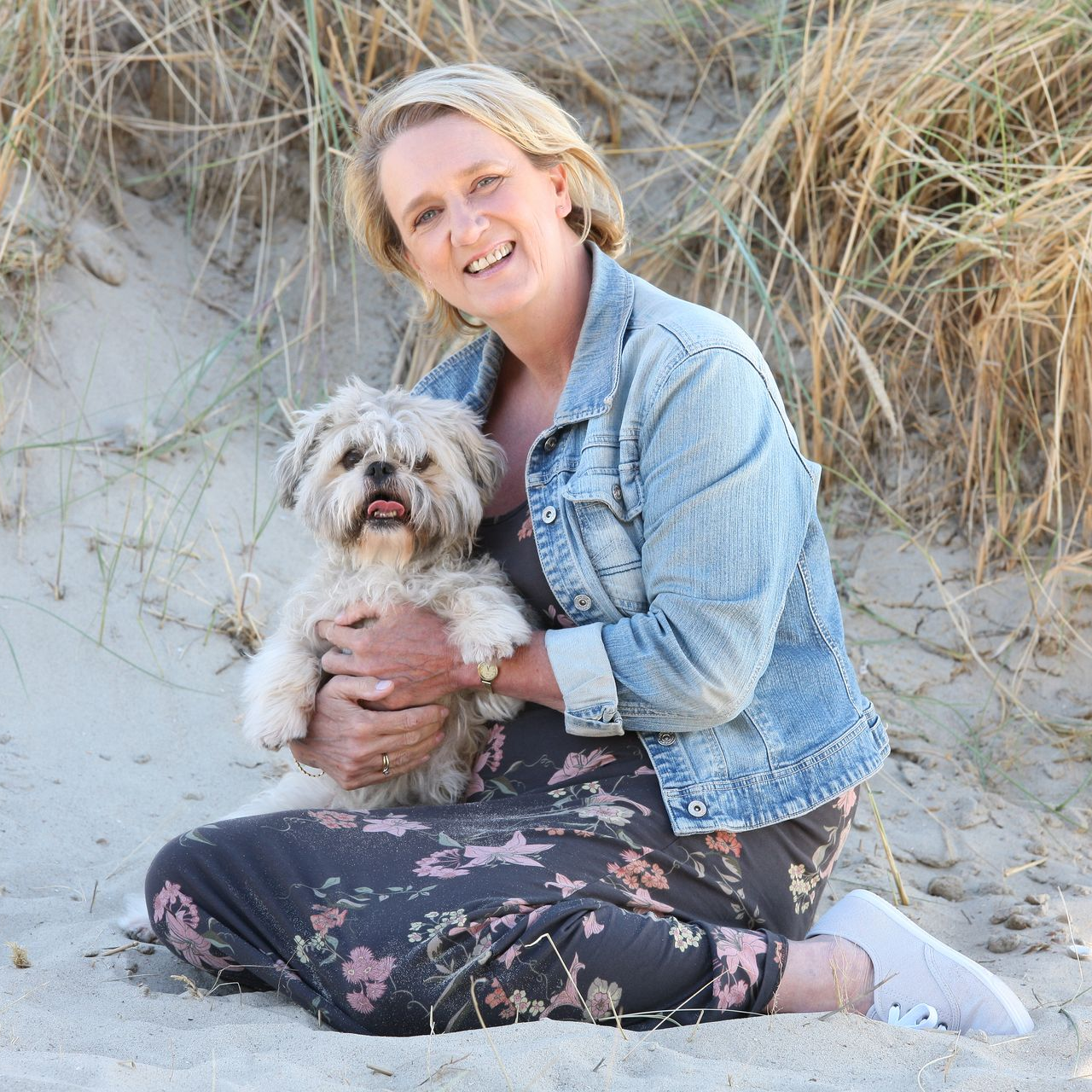 Bibian in spijkerjasje, bloemenjurk en sneakers, zittend op het zand met  hondje Binky in haar armen; strandsetting