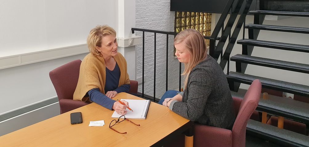 Twee vrouwen zitten aan een tafel, één schrijft op een papier. Op de achtergrond is een trap te zien.