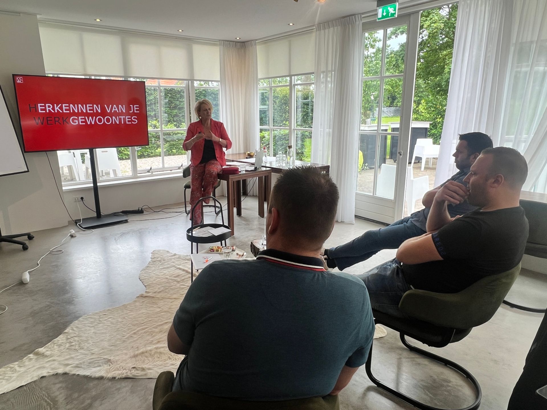 Een vrouw presenteert aan drie mannen in een goed verlichte kamer. Er zijn een rood scherm, witte gordijnen en een bontkleed zichtbaar.