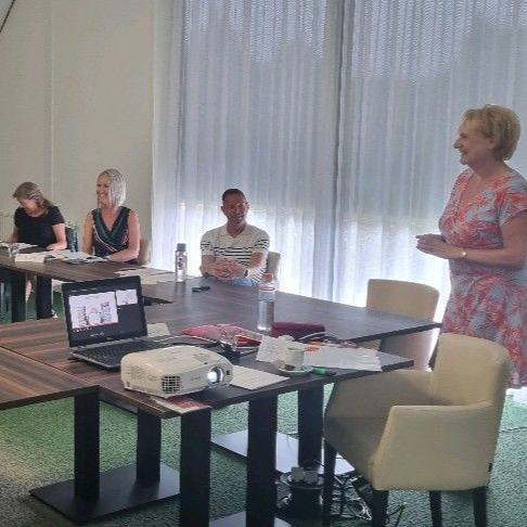 Mensen in een vergadering, een vrouw die een presentatie geeft. Tafels, een laptop met projector, waterflessen. Een lichte ruimte met gordijnen.