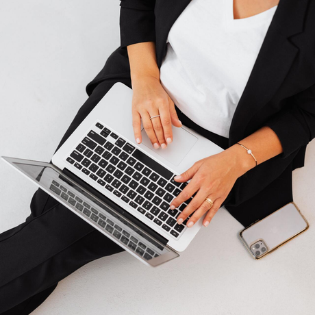 Vrouw in zwart pak zit met laptop en telefoon op wit oppervlak.