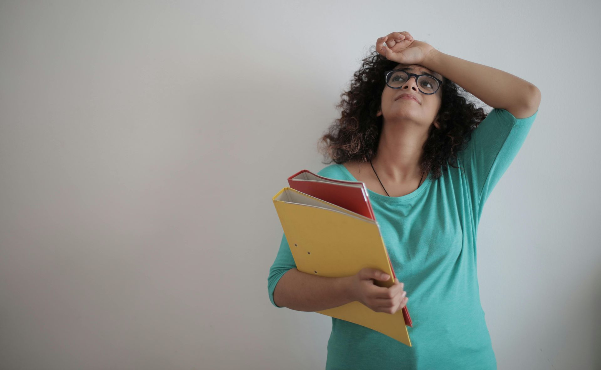 Vrouw met krullend haar en een bril, die dossiers vasthoudt, kijkt overweldigd omhoog met haar hand op haar voorhoofd.