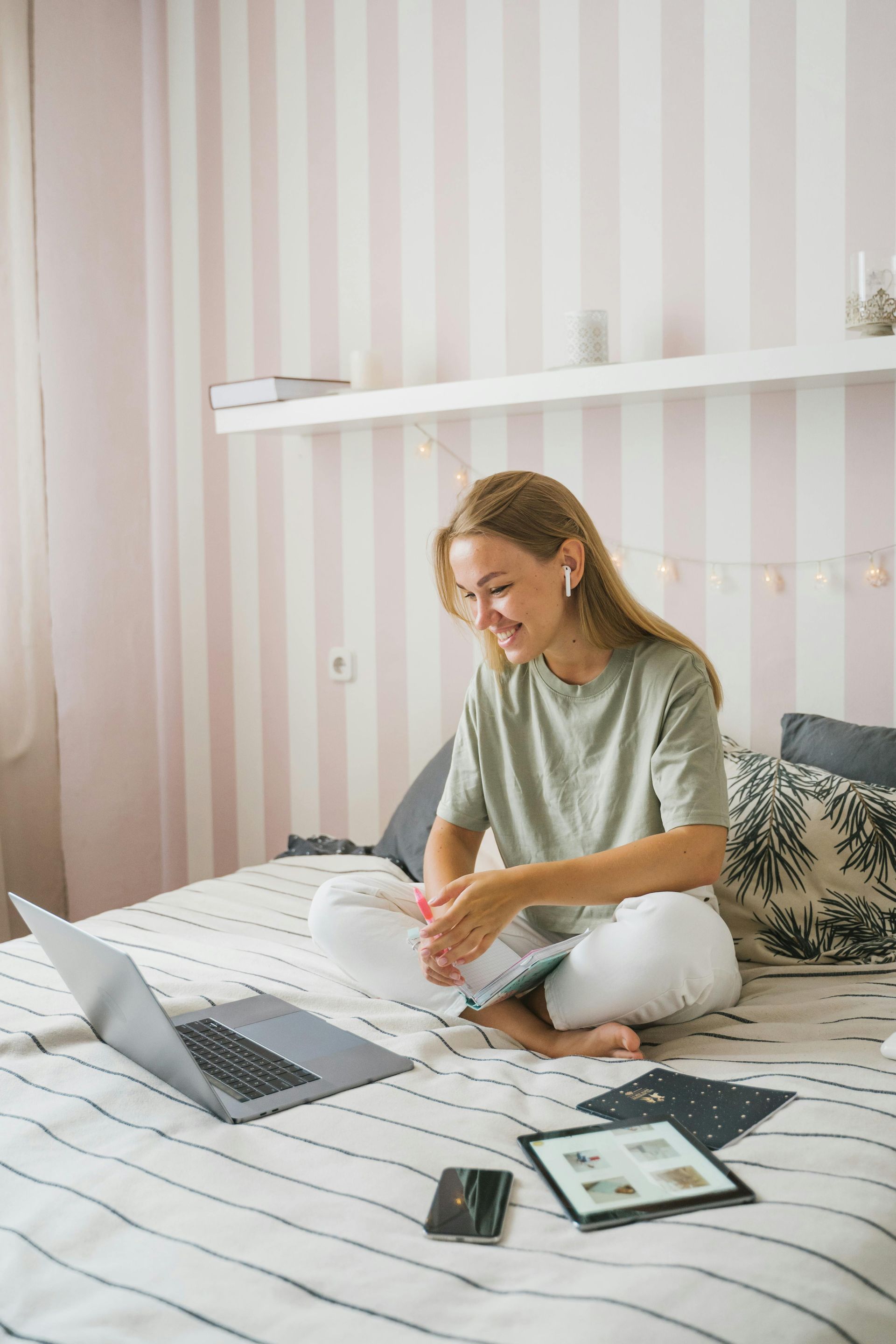 Een vrouw zit met gekruiste benen op bed en glimlacht naar een laptop. Ze draagt ​​een groen shirt en een witte broek.