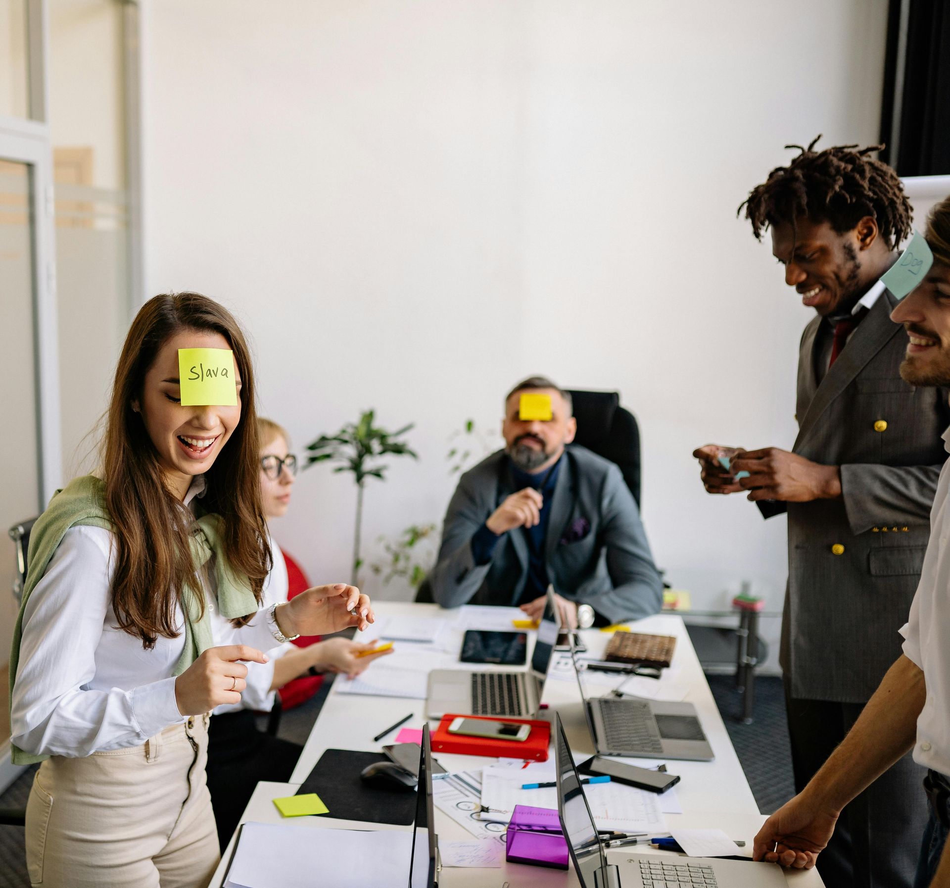 Een team van medewerkers speelt een woordspel, lachend en met plakbriefjes op hun voorhoofd, rond een tafel met laptops.