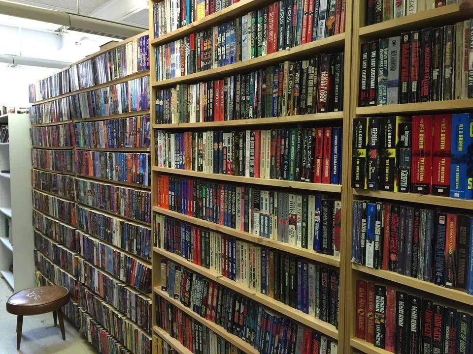 Bookshelves filled with books in a library or bookstore. A small stool is in the corner.