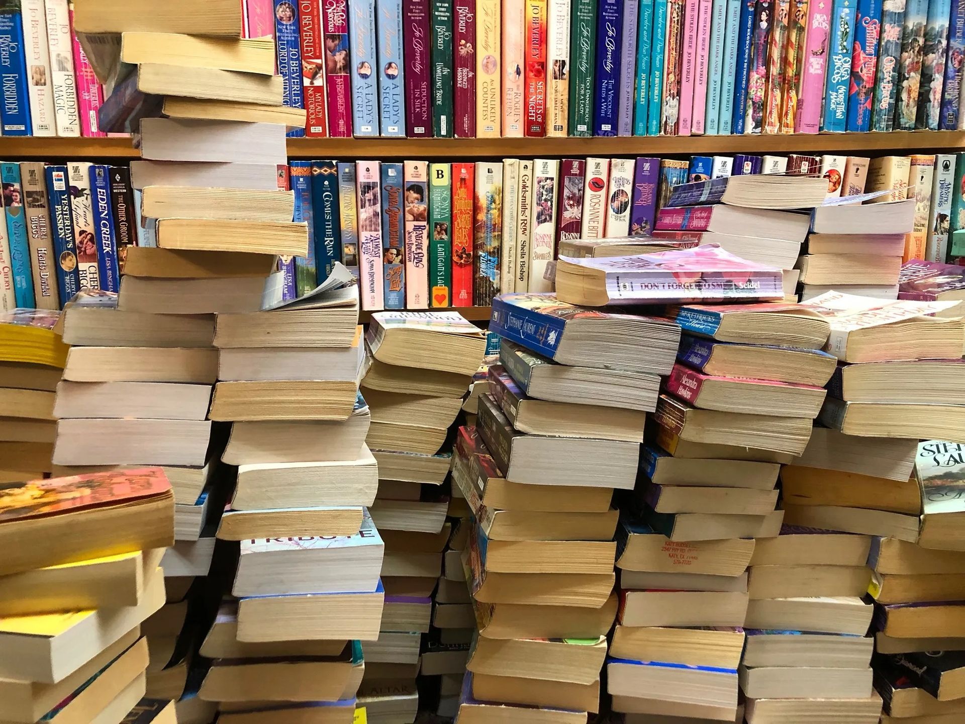Piles of stacked books in a used bookstore, shelves of colorful books in the background.