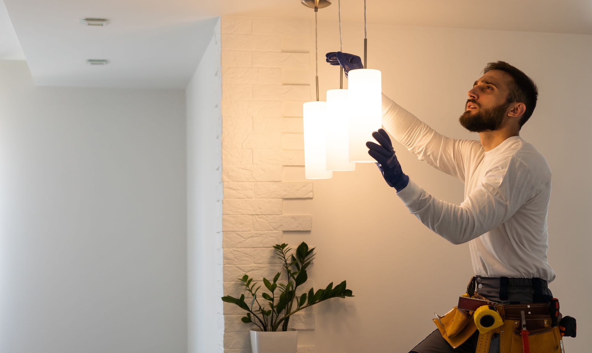 Man in gloves installing a modern light fixture. He's wearing a tool belt and looking up.