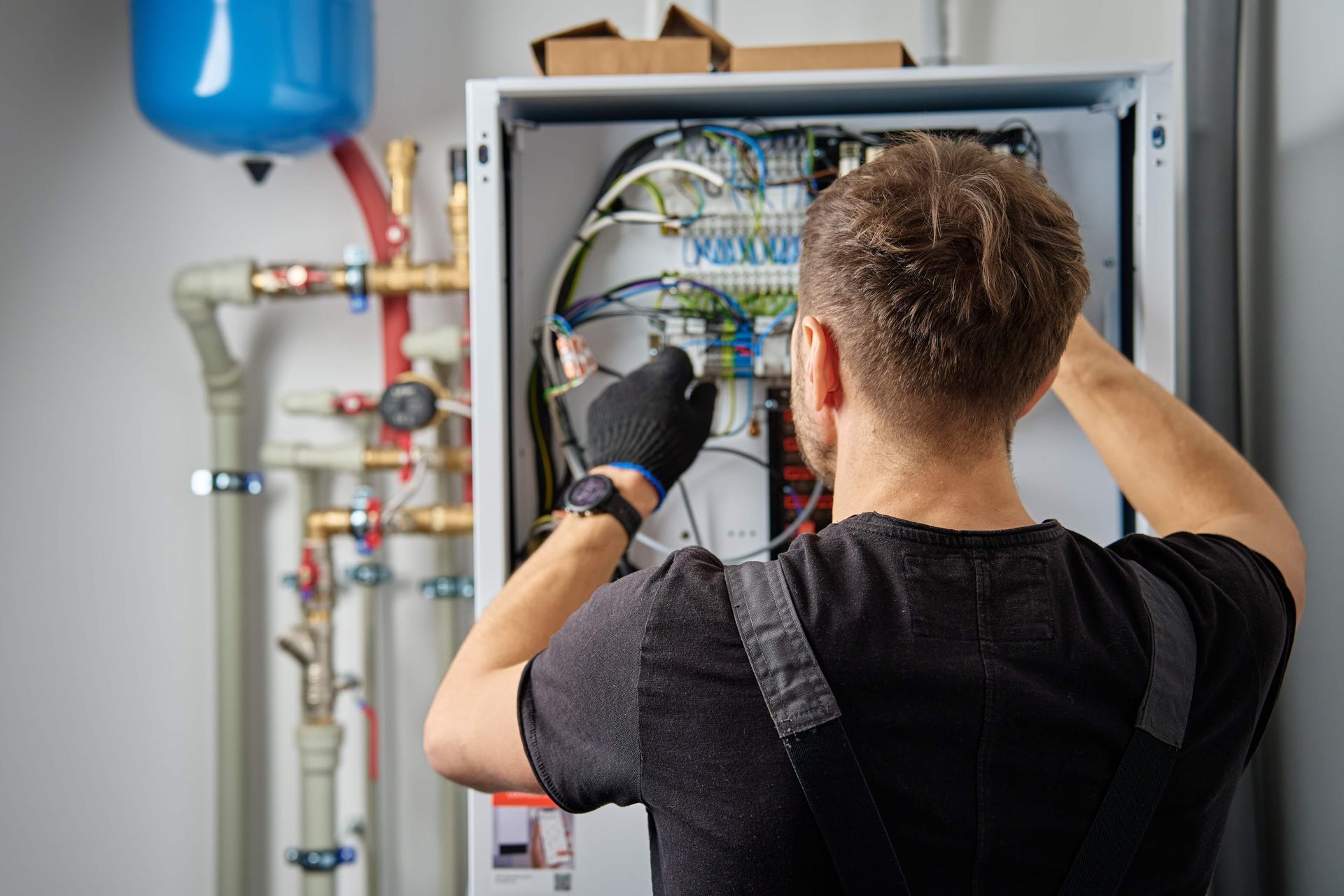 Electrician working on wiring inside a white electrical panel, near pipes and a blue tank.