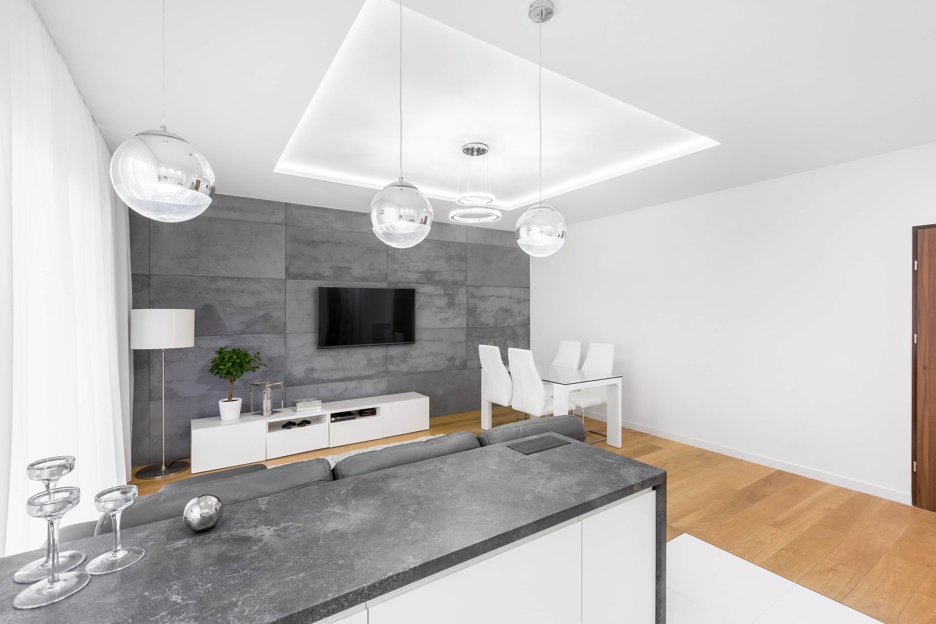 Modern living room with gray accent wall, TV, white furniture, and wood floors.