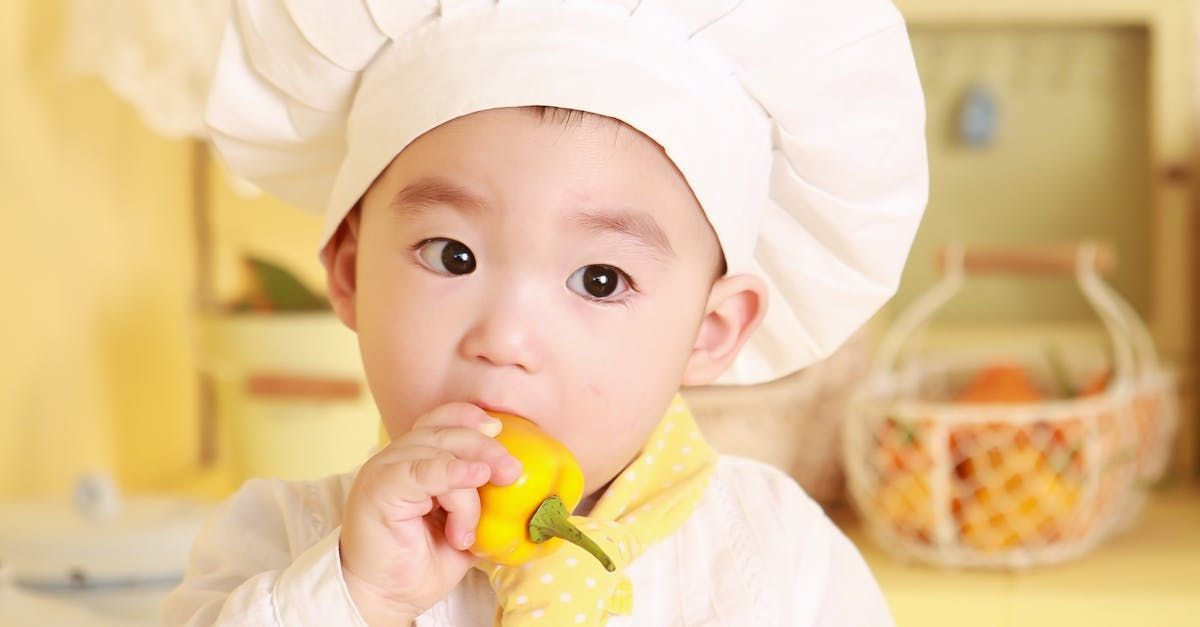 A baby wearing a chef 's hat is eating a yellow pepper.