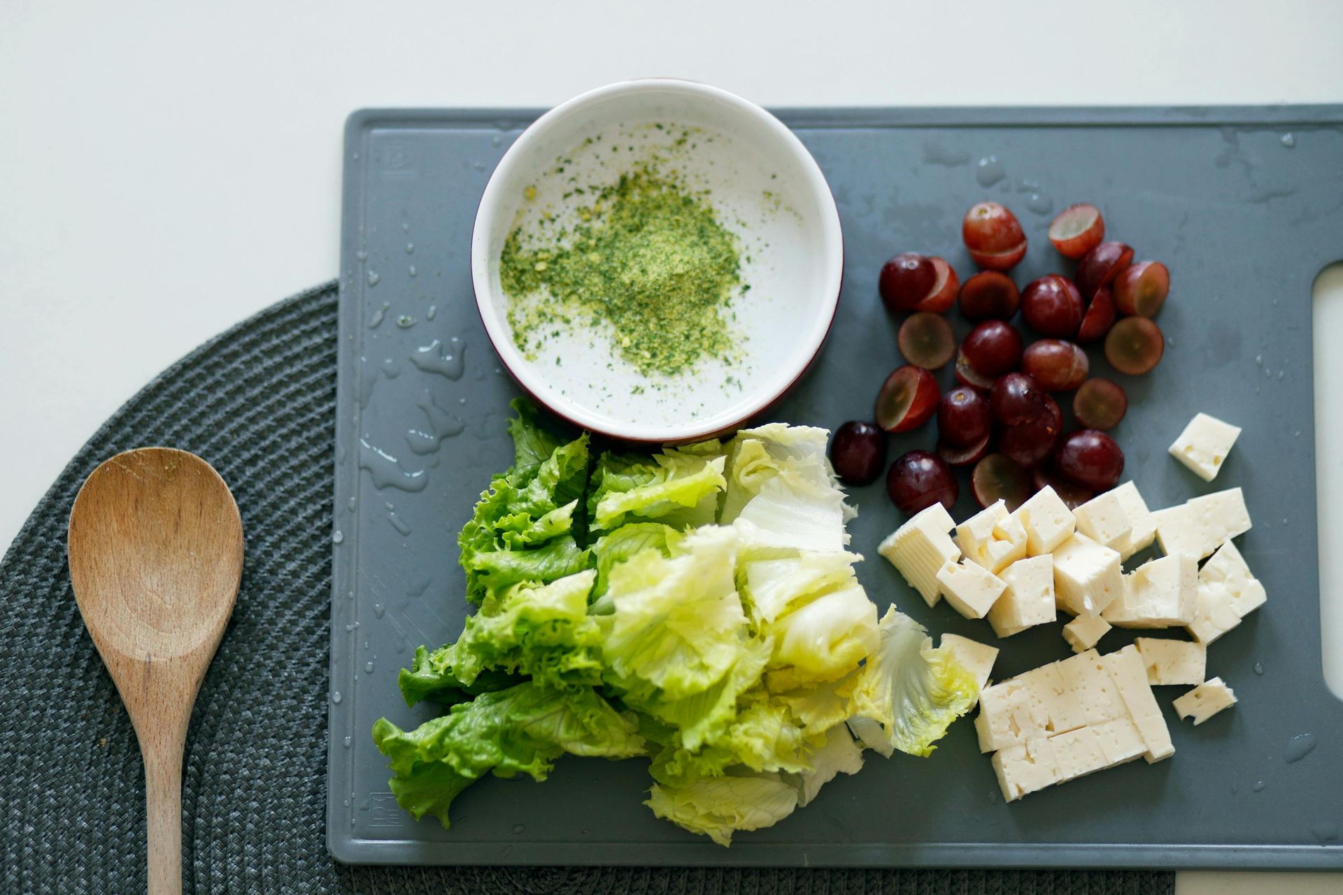 A cutting board with lettuce , grapes , cheese and a bowl of green powder.