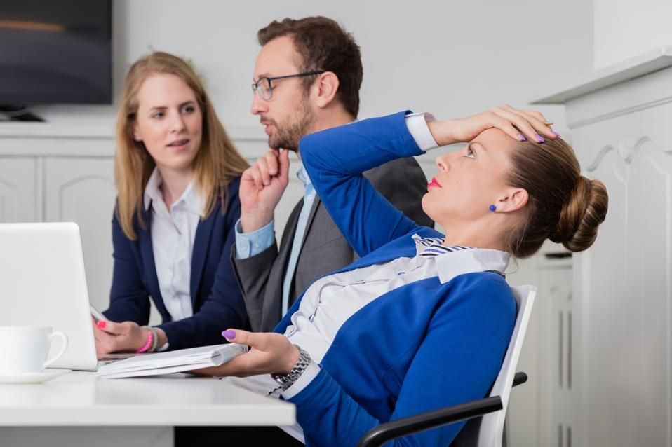 Three office workers at a desk, with one female hitting their forehead in disappointment.
