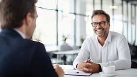 Two men in a bright office meeting, one smiling across a table with coffee cups and papers.