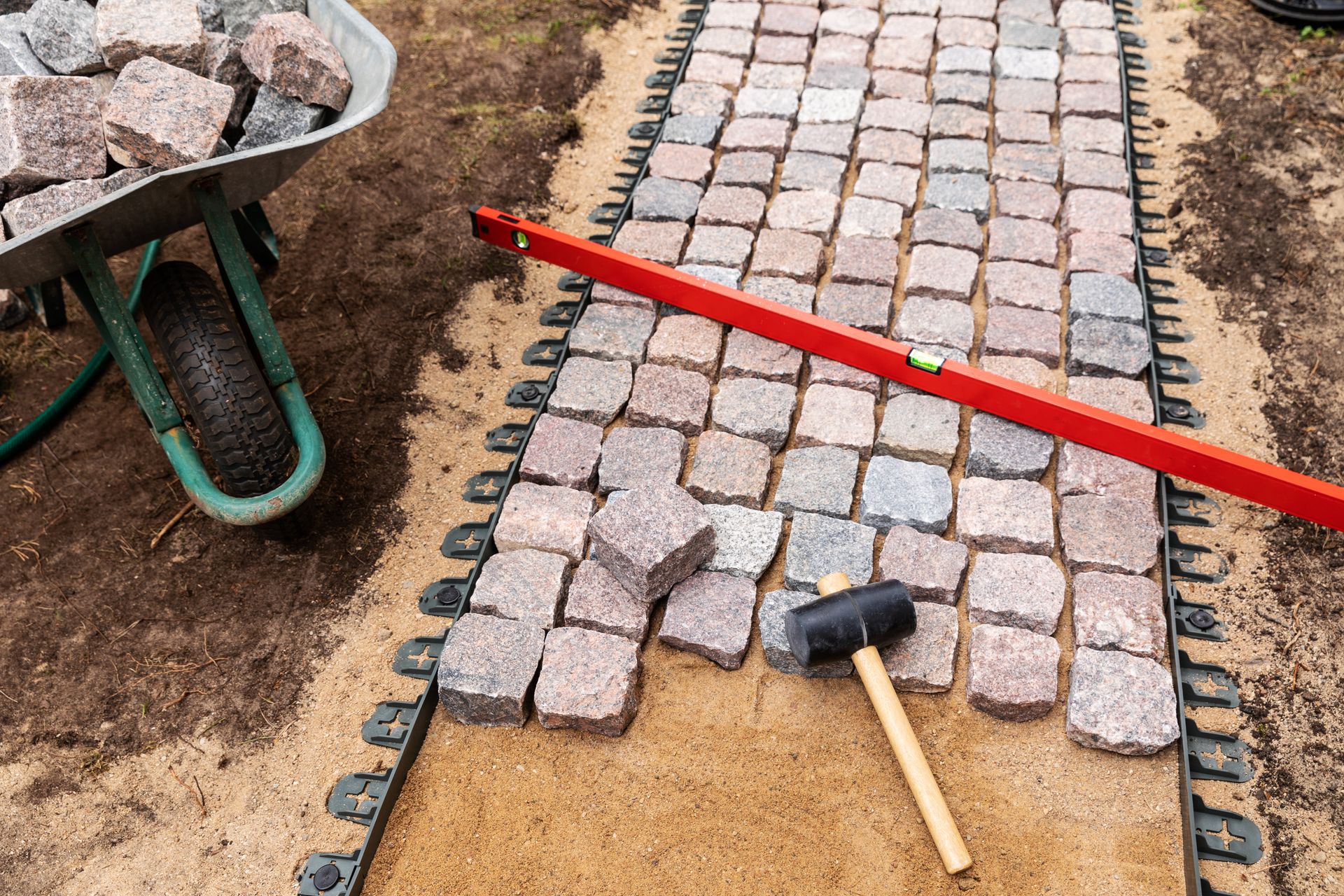 Pathway construction: granite pavers laid on sand base, with a level and rubber mallet. Wheelbarrow of stones in the background.