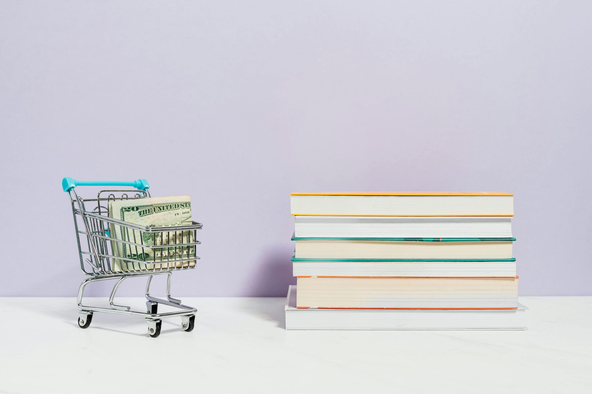 A miniature shopping cart filled with dollar bills sitting next to a stack of books against a light purple background.