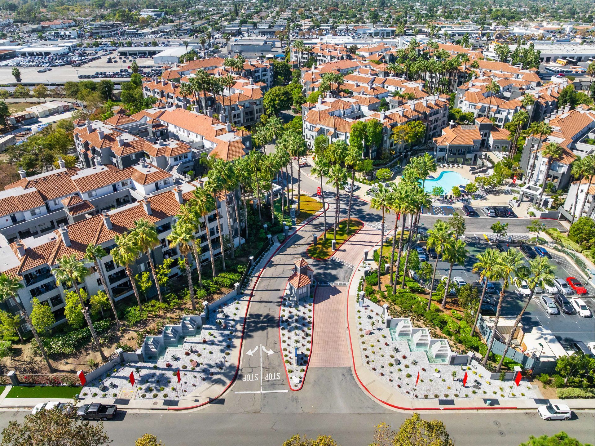 Aerial view of an apartment complex with red-tiled roofs, palm trees, and a pool. Entry road is marked.