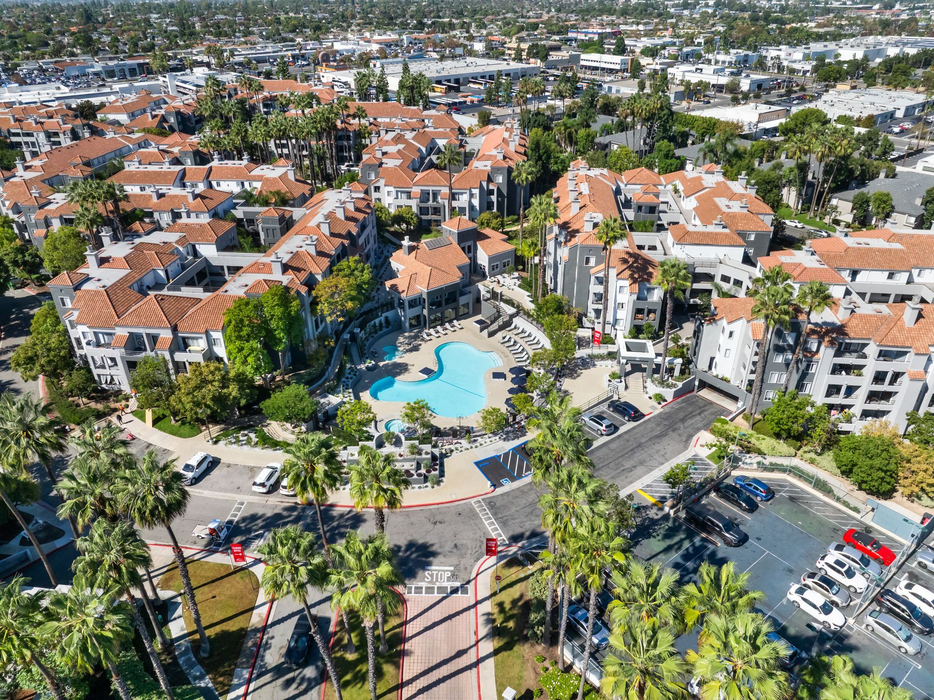 Aerial view of a residential complex with a central swimming pool and palm trees. Red-tiled roofs and parking areas.