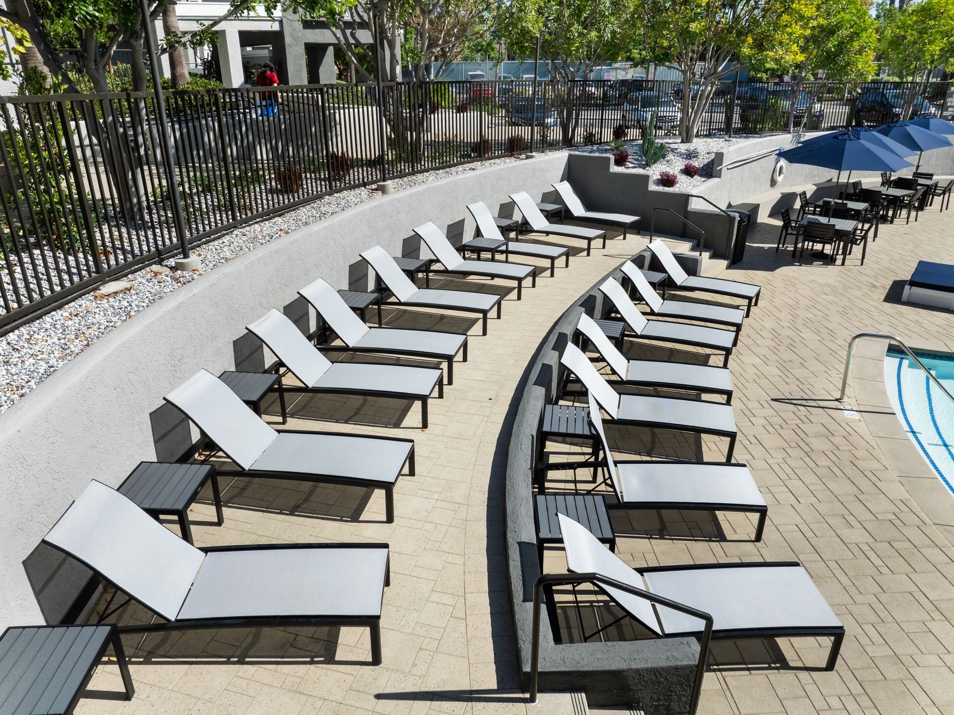 Poolside lounge chairs with side tables arranged in a curved row, with a black fence and part of a pool visible.