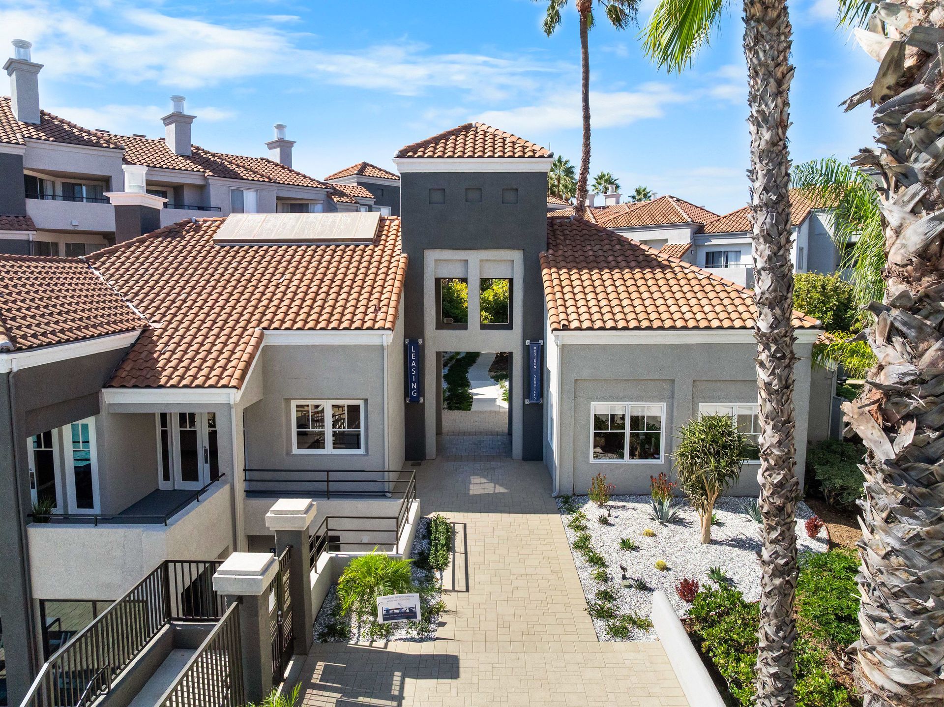 Building with terracotta roof tiles, gray walls, and pathway lined with landscaping.