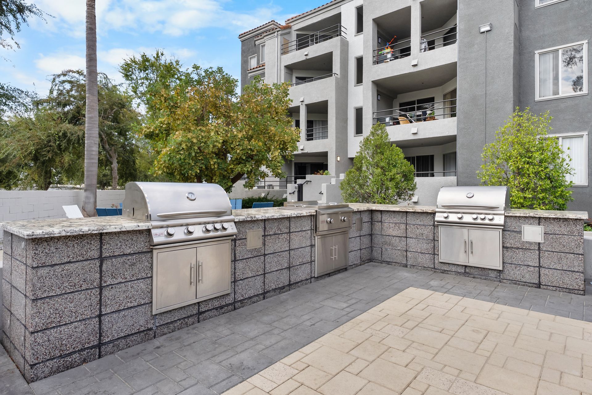 Outdoor grilling station with stainless steel grills and gray stone countertops in front of an apartment building.
