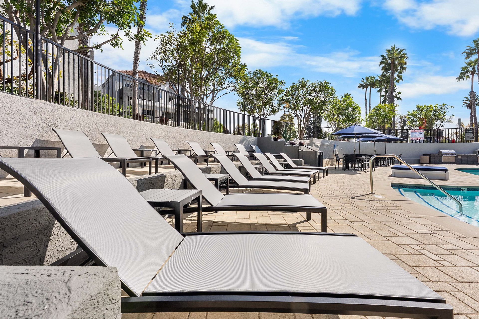 Lounge chairs by a pool on a sunny day. Trees and buildings in the background.