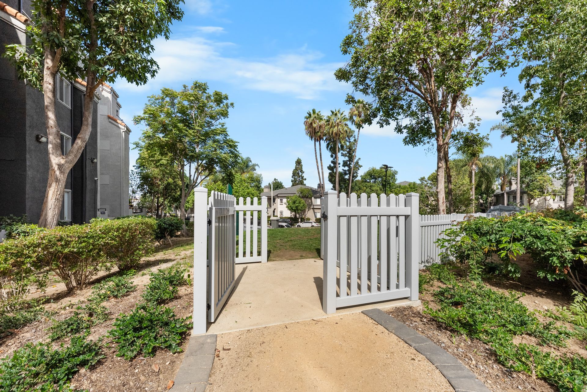 Gray picket fence with open gate, pathway, and landscaping leading to a green space dog park.