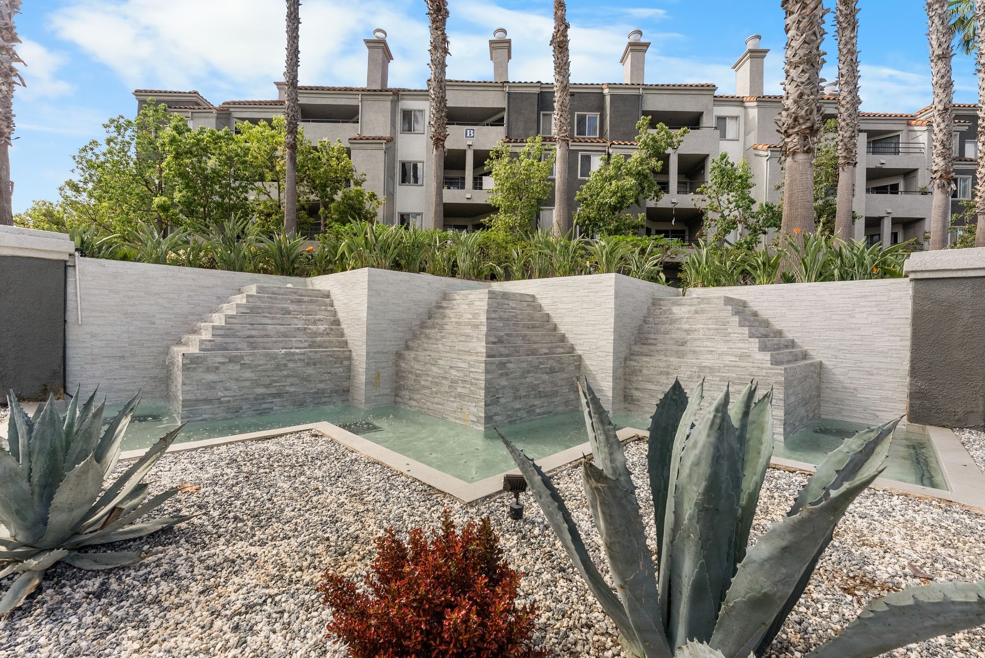 Water fountain with layered stone, agave plants, and apartment building in background.