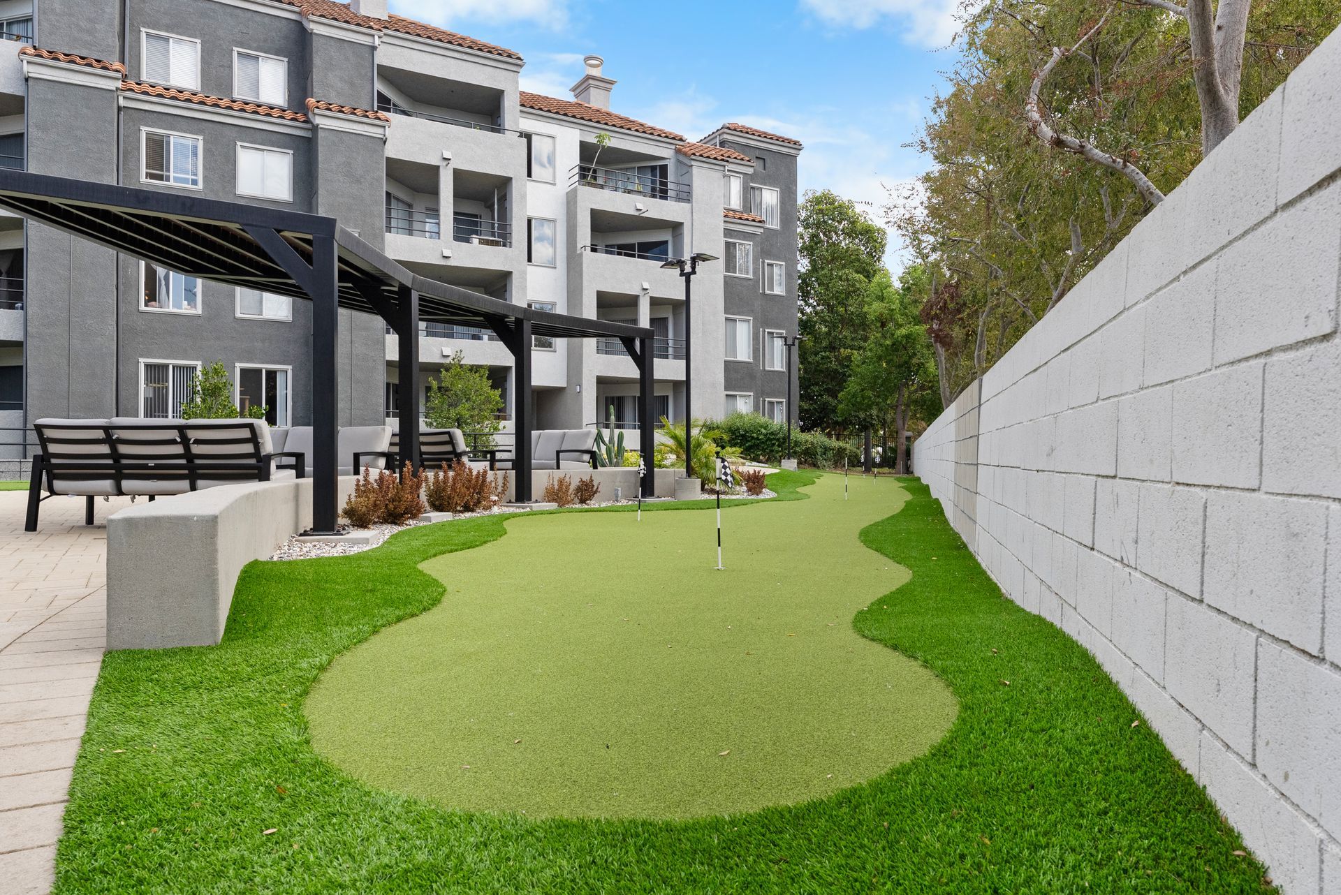 Putting green with flag in front of gray apartment building, surrounded by lawn and a stucco wall.