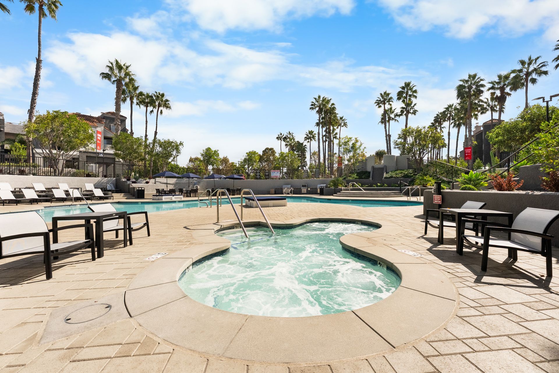 Pool area with a spa, lounge chairs, tables, and palm trees under a blue sky.