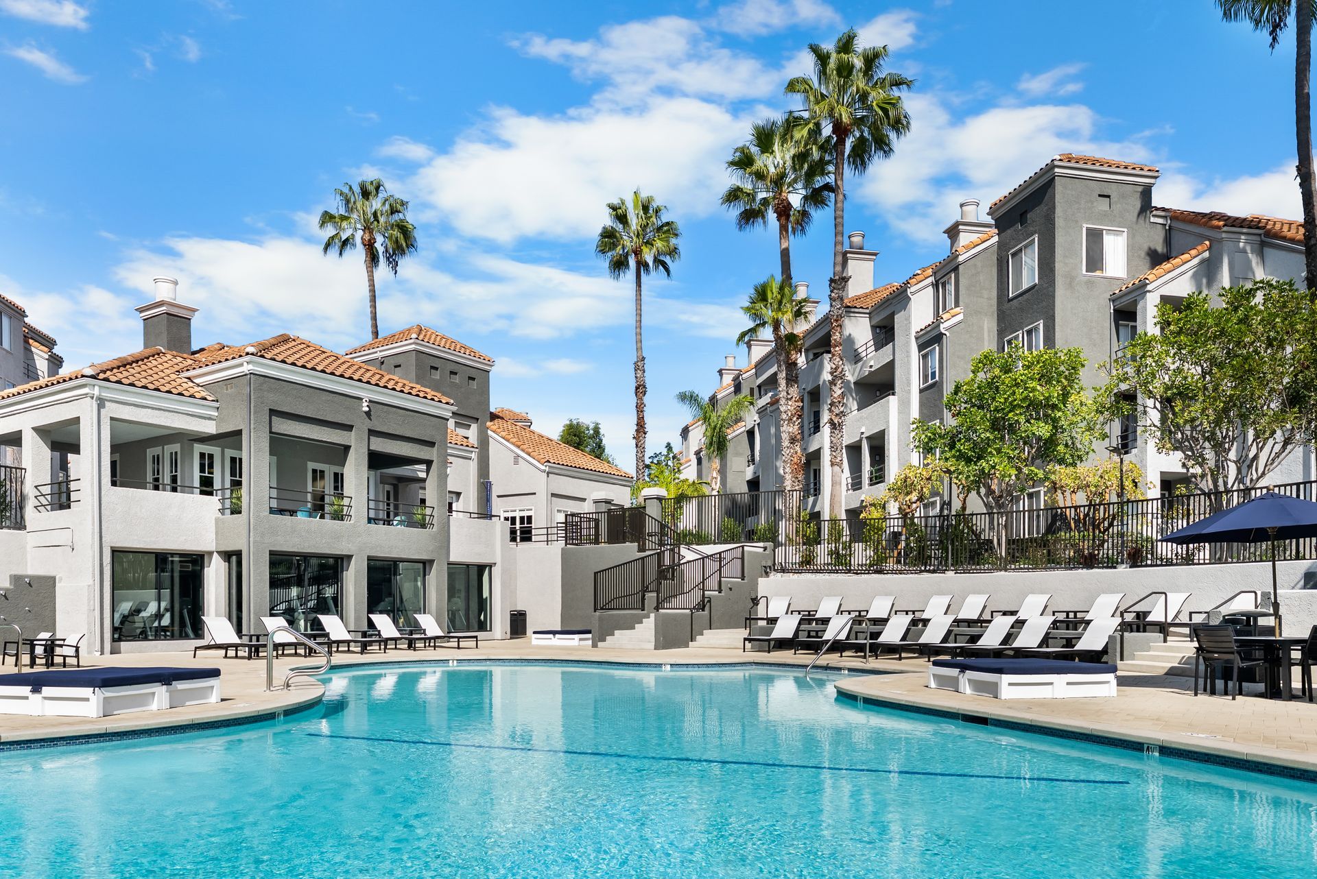 Swimming pool in front of apartment buildings with palm trees and lounge chairs on a sunny day.