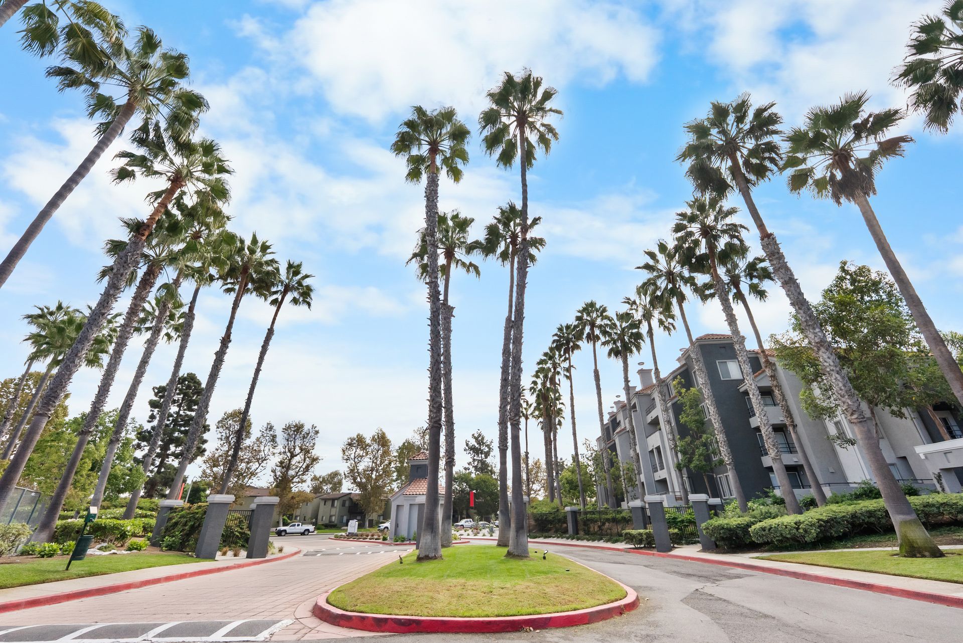 Palm trees line a street leading to a gated apartment complex on a sunny day.