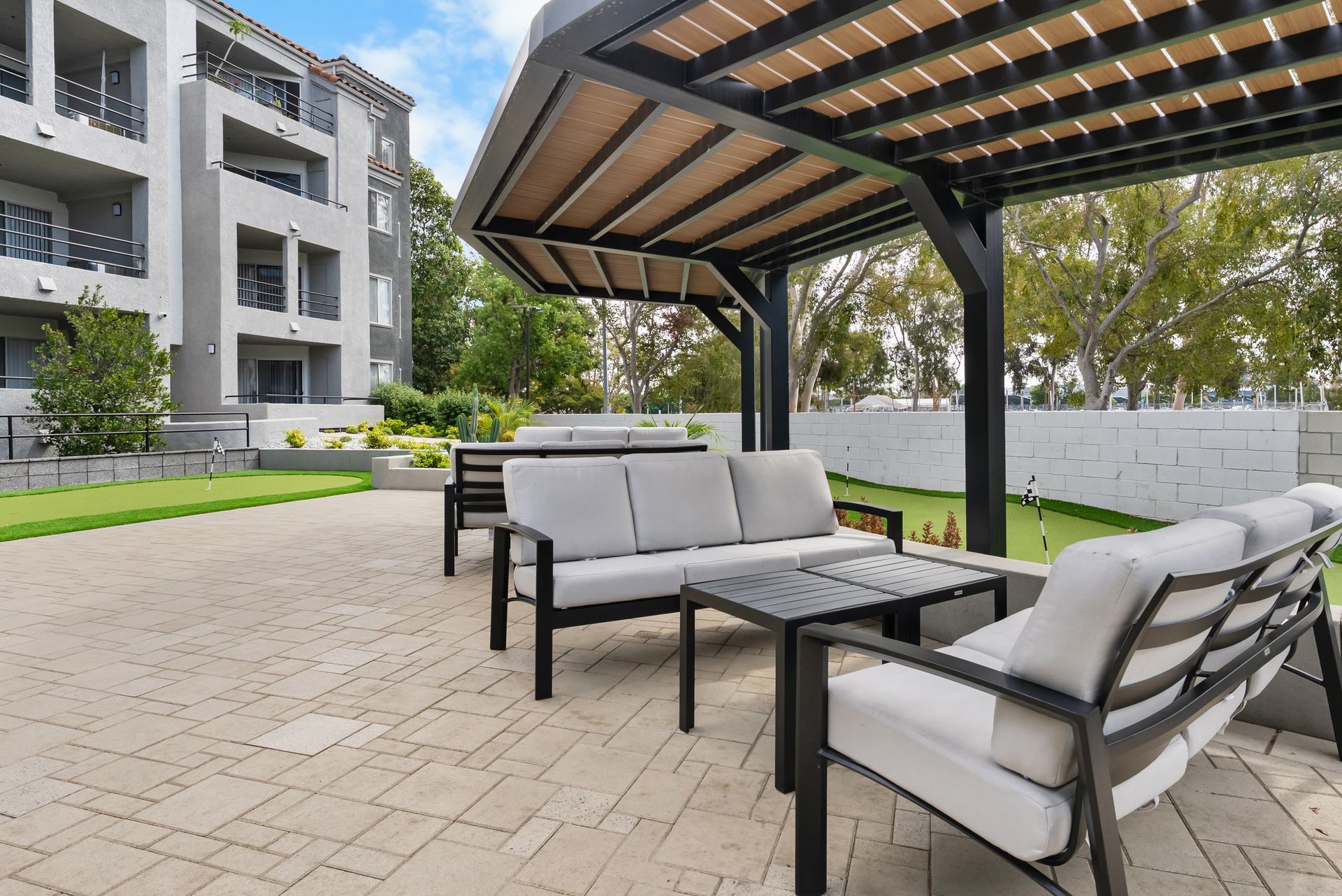 Patio seating under a pergola with a building in the background. Gray furniture on a stone patio.