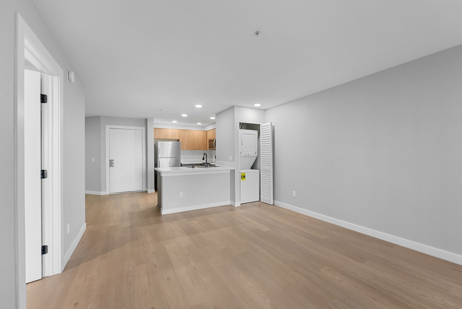 An empty living room with hardwood floors and a kitchen in the background.