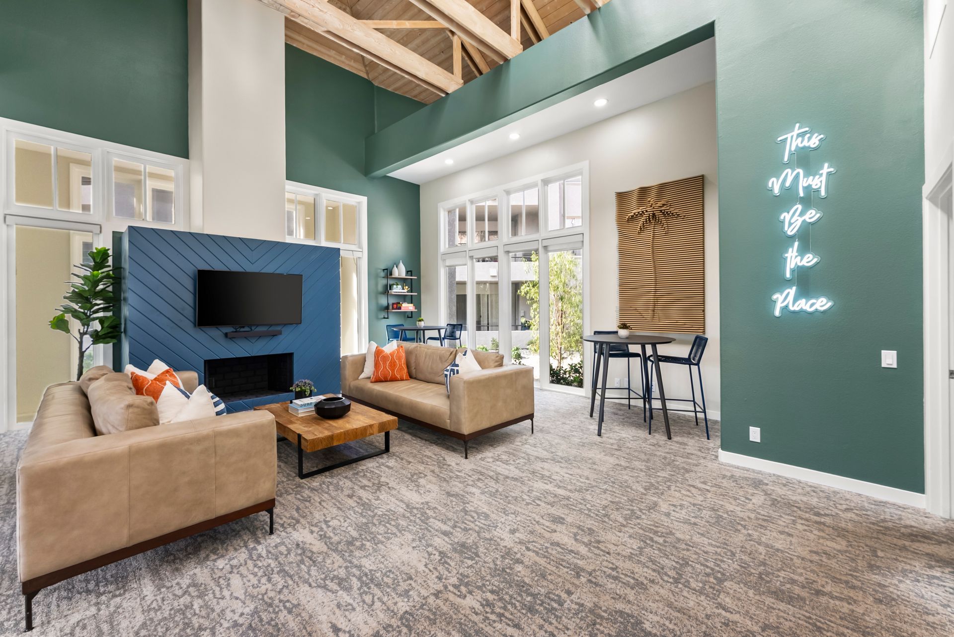 Lounge with beige sofas, blue accent wall with TV, fireplace, and neon sign. Windows to the outdoors.