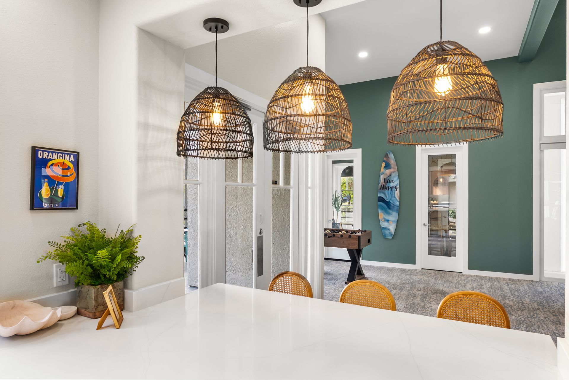 Three woven pendant lights over a white countertop, leading into a green-walled room with a surfboard and doorway.