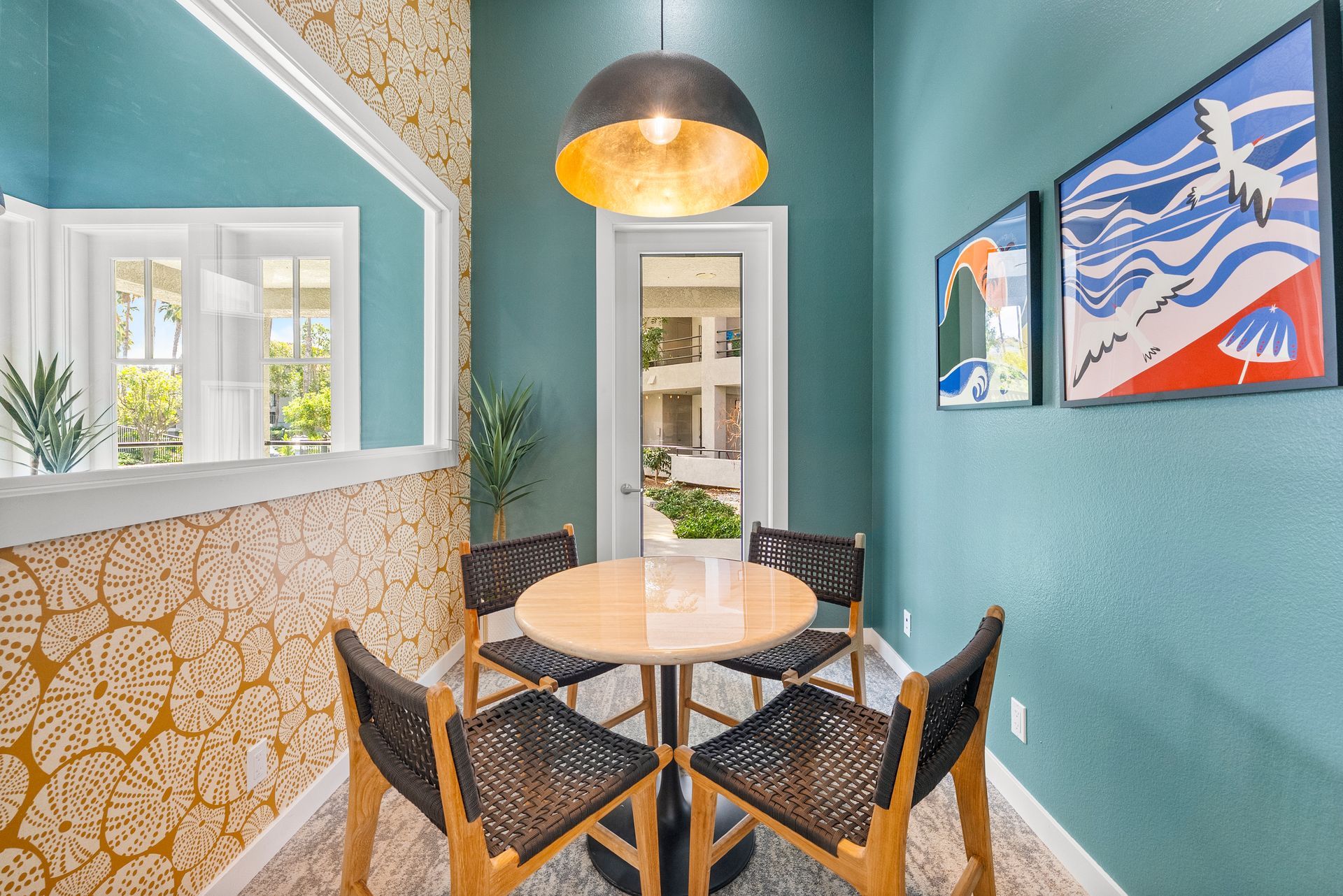 Small dining nook with round table and four chairs. Teal walls with artwork; gold pendant light.