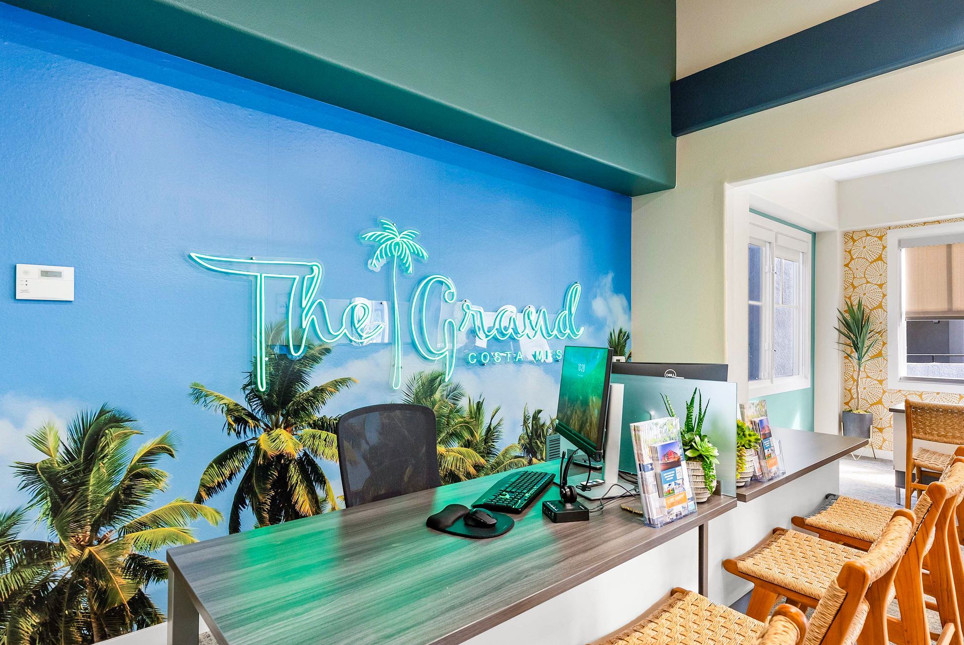 Reception desk with a tropical mural and neon sign; several woven barstools and a computer on the desk.