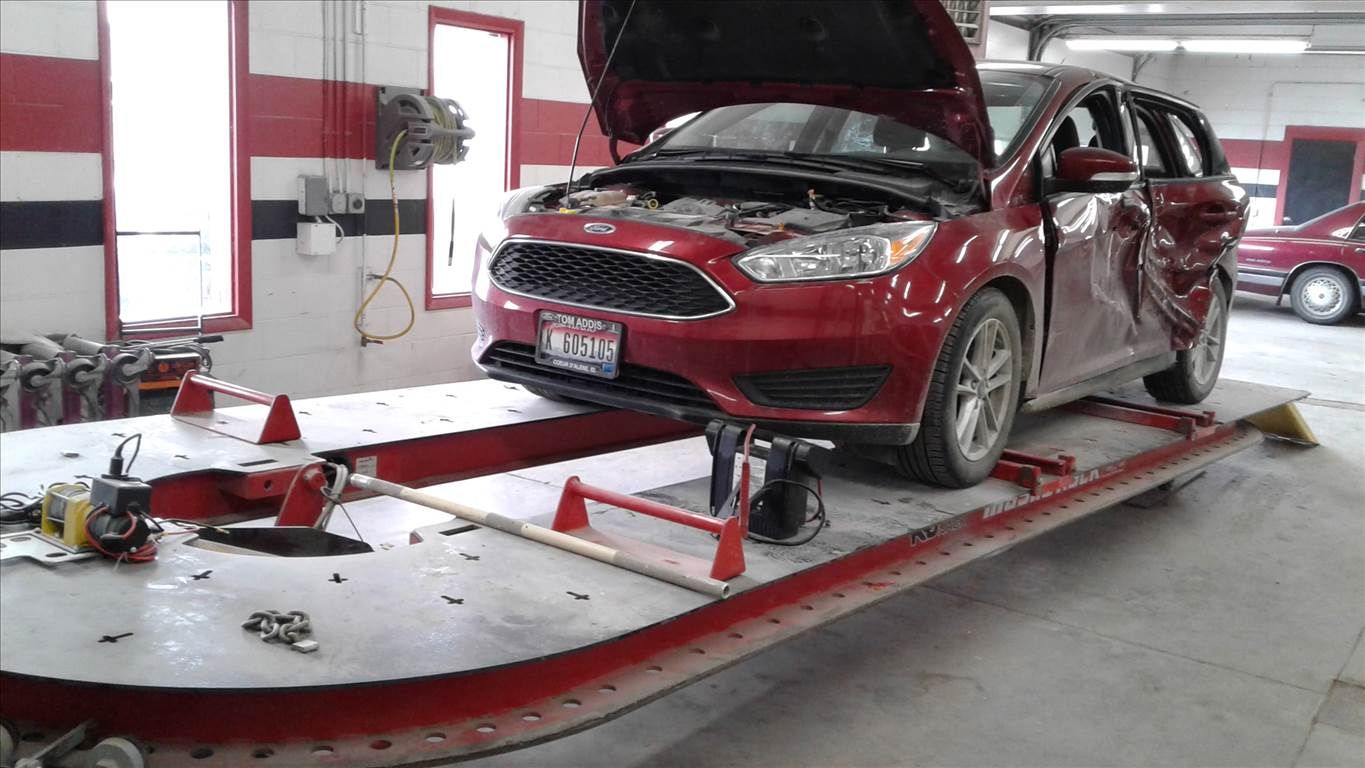 Red car on a vehicle lift in a garage, with the hood open and front damage visible.