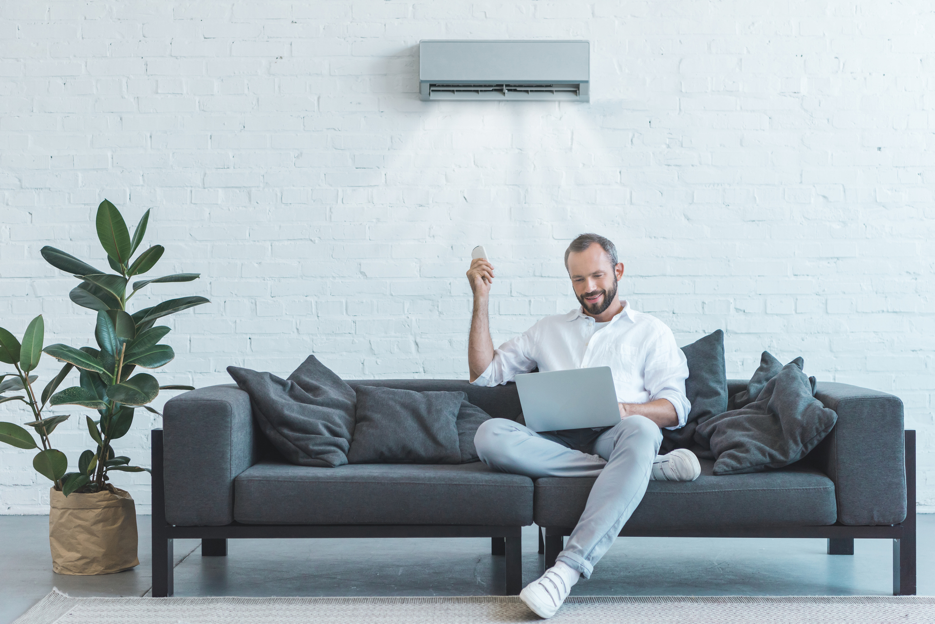Man on couch using laptop, holding remote, air conditioner above, white brick wall.