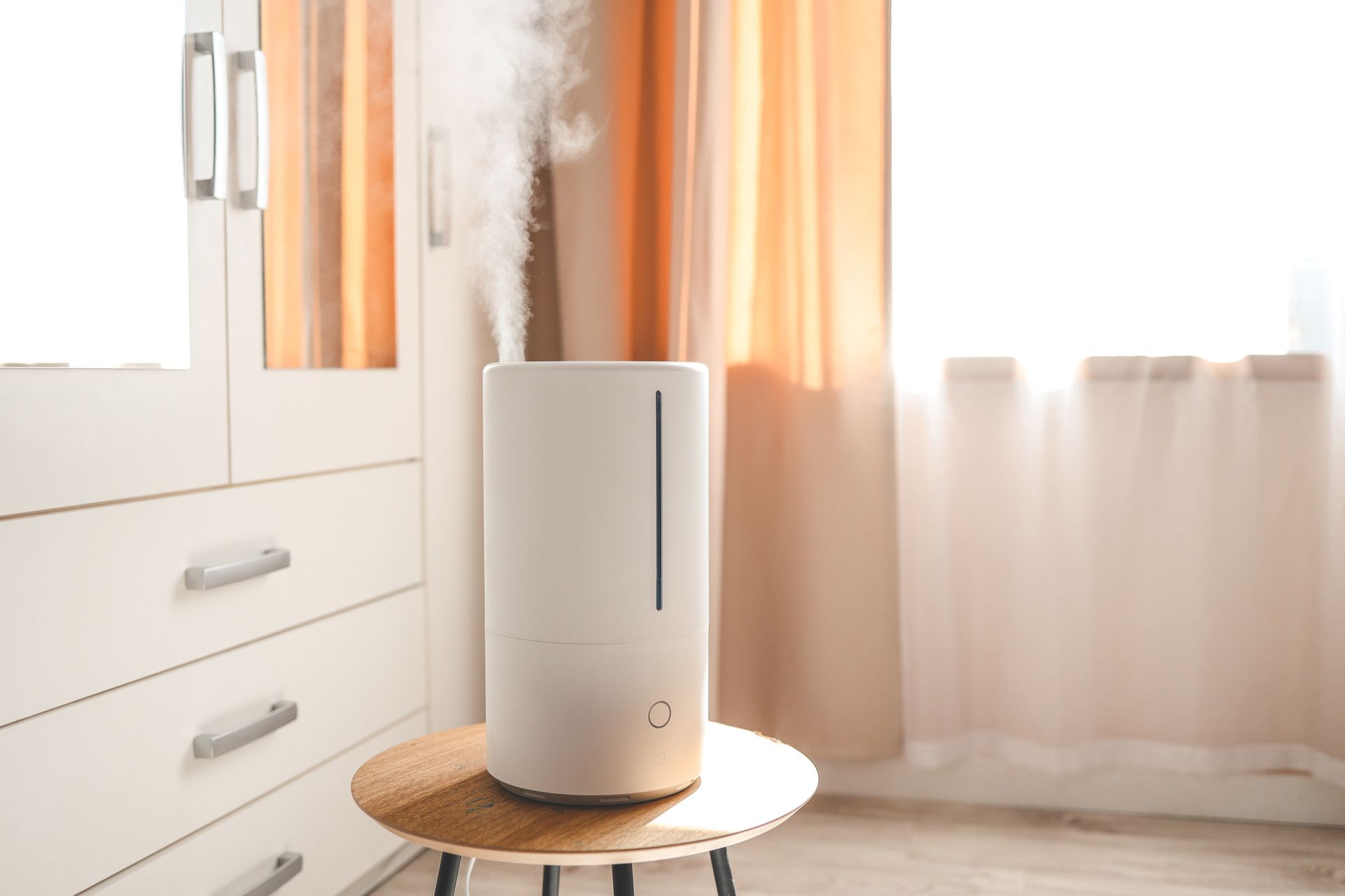 White humidifier on a small table, emitting steam in front of a window with sheer curtains.