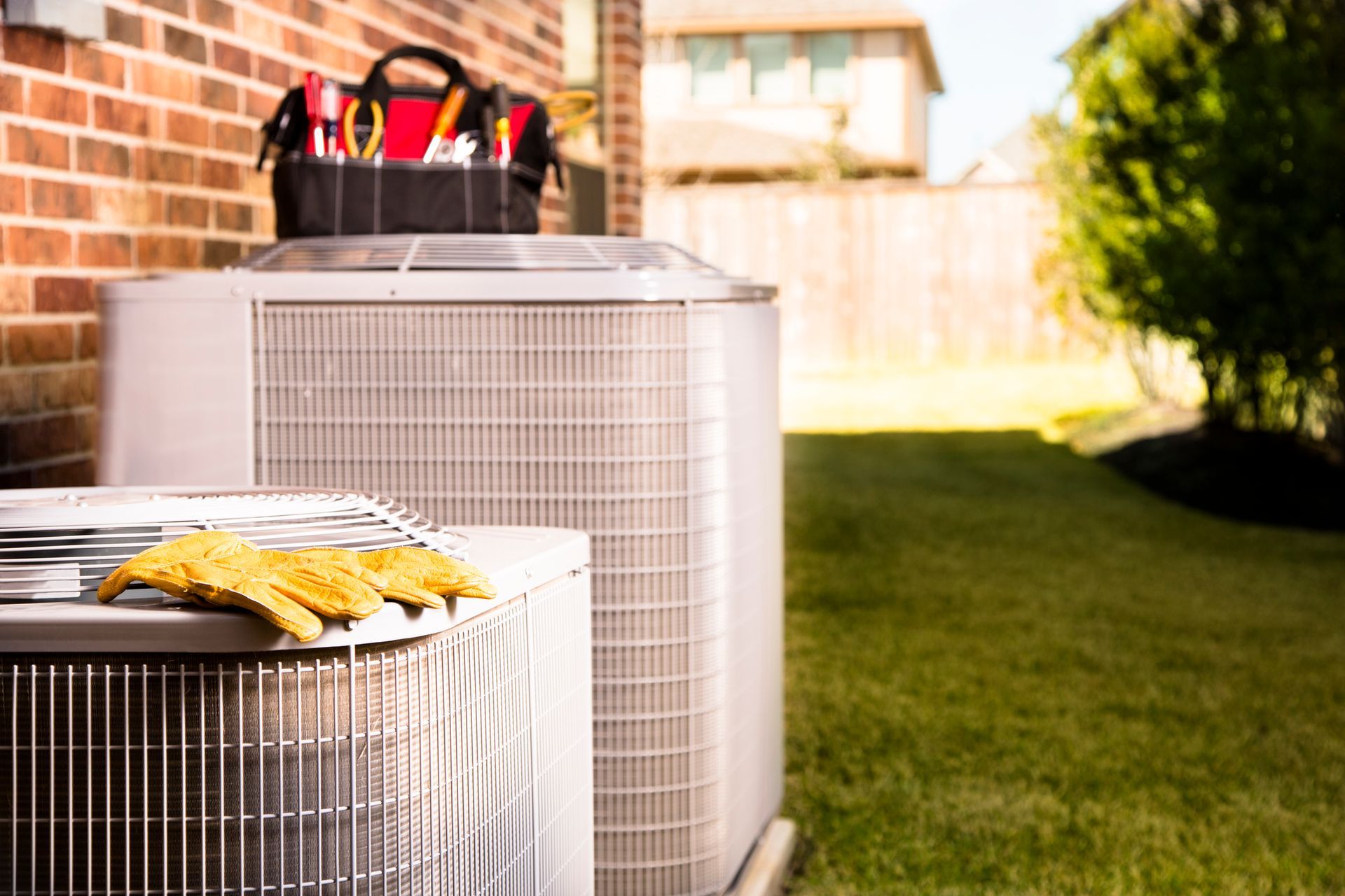 Two air conditioning units in a yard; toolbox on top, work gloves on one unit.