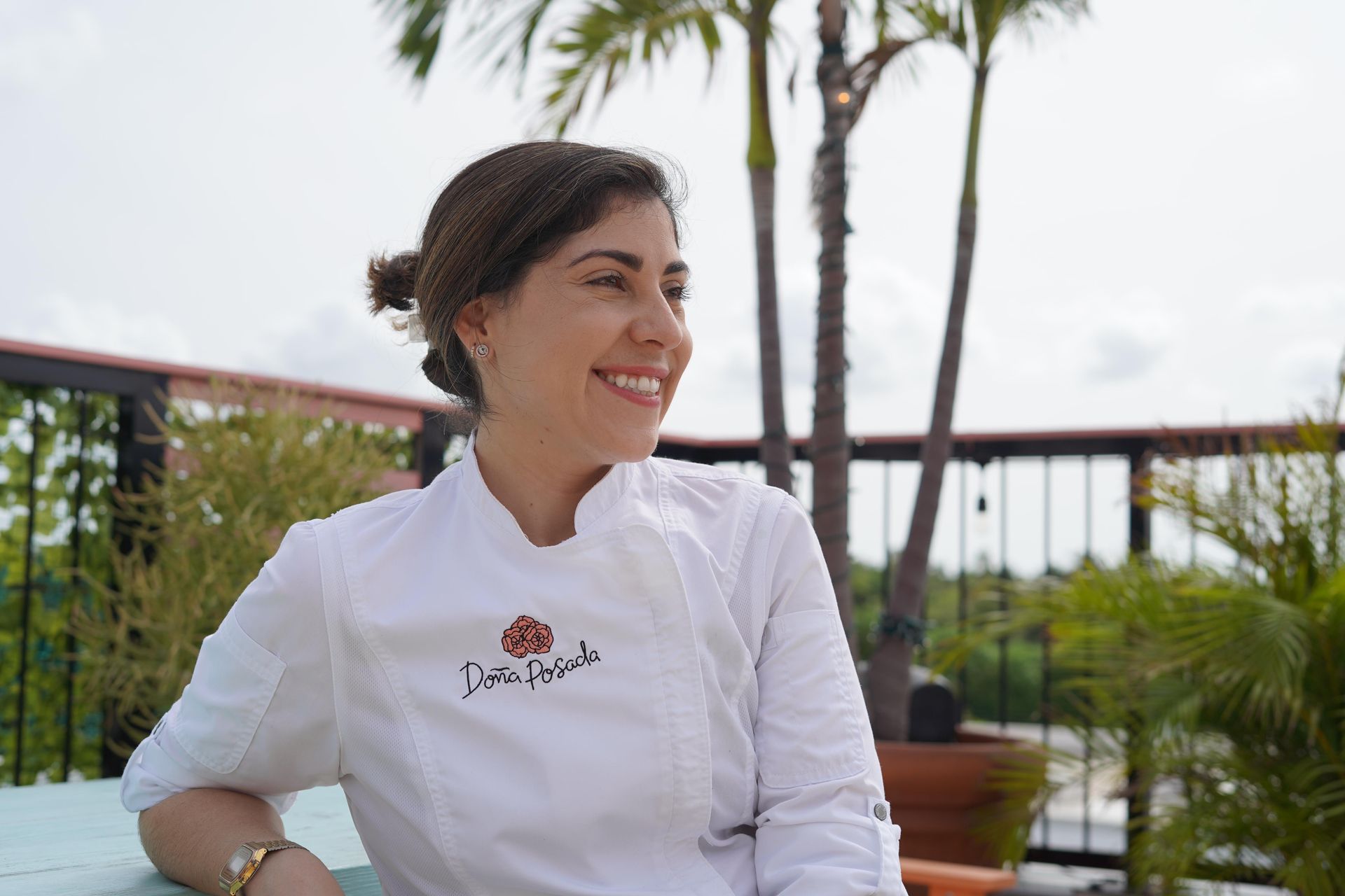 michele cruz in a white chef 's jacket is smiling while sitting at a table.