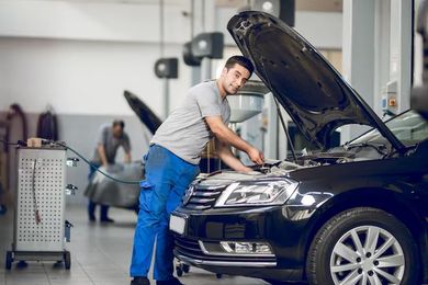 A mechanic in a grey shirt and blue pants works on the engine of a black car in an auto repair shop.