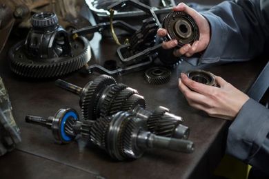 A person in a workshop disassembles mechanical parts, including various metal gears and shafts laid out on a table.
