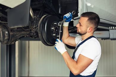 A mechanic wearing gloves and overalls uses a wrench to work on a car's brake drum in an auto repair shop.